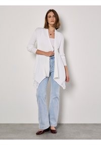 Witte geribbelde cardigan met lange mouwen en open voorkant, voorzien van kant aan de onderkant, gecombineerd met lichtblauwe flared jeans en bordeauxrode ballerina's.