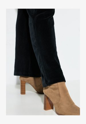 Persona vestida con pantalones de pana negros y botas de tacón alto de ante color beige con tacones de bloque de madera, de pie sobre un suelo blanco.