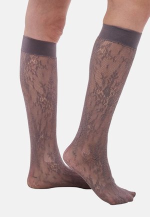 Gambe che indossano calze autoreggenti trasparenti in pizzo con motivo floreale e bande elastiche solide nella parte superiore su sfondo bianco.