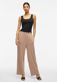 Top negro sin mangas combinado con pantalones anchos beiges, holgados, que cuentan con una cinturilla elástica y bolsillos laterales. La modelo lleva sandalias de tacón negras.