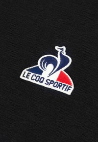 Tessuto nero con un logo ricamato in bianco, blu e rosso che presenta un gallo stilizzato e il testo "LE COQ SPORTIF" sottostante.