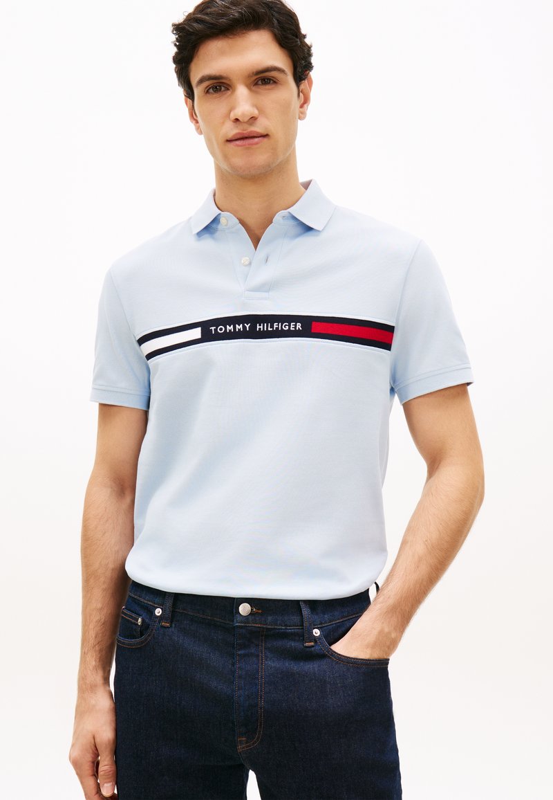 Άνδρας που φοράει ανοιχτό μπλε πόλο Tommy Hilfiger με το λογότυπο και κόκκινη, λευκή και μαύρη ρίγα στο στήθος, σκούρο τζιν, με το χέρι στην τσέπη.