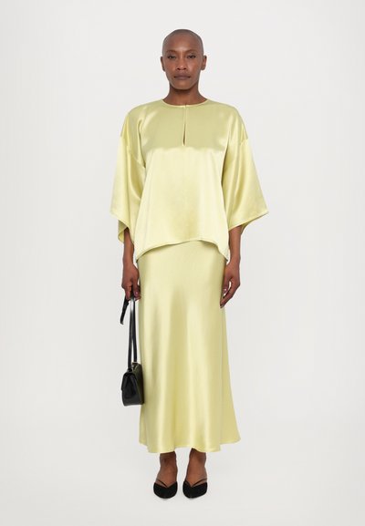 Top de satén amarillo claro con escote redondo y mangas anchas, combinado con una falda larga a juego. La modelo sostiene un bolso negro.