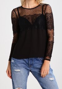 Schwarzes Langarmshirt aus transparentem gepunktetem Mesh mit Spitze. Rundhalsausschnitt und entspannter Schnitt, kombiniert mit zerrissenen blauen Jeans.