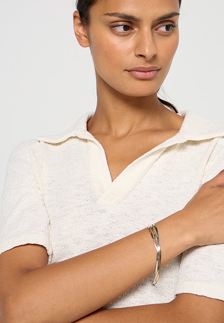 Krótki rękaw, biały, dziany top z kołnierzykiem polo; modelka nosi srebrną skręconą bransoletkę na nadgarstku. Prosta i fakturowana tkanina.