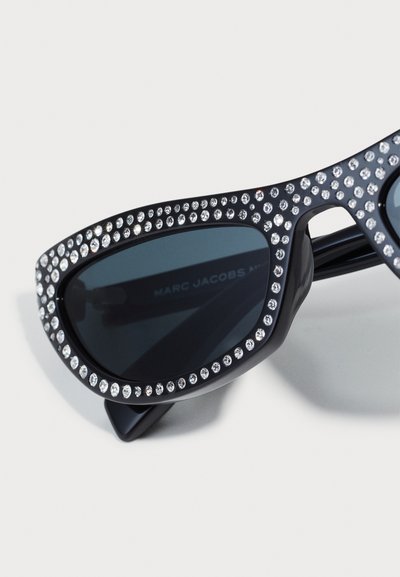 Lunettes de soleil de style œil de chat avec une monture noire ornée de strass transparents le long du bord supérieur, dotées de verres foncés et de branches de marque.