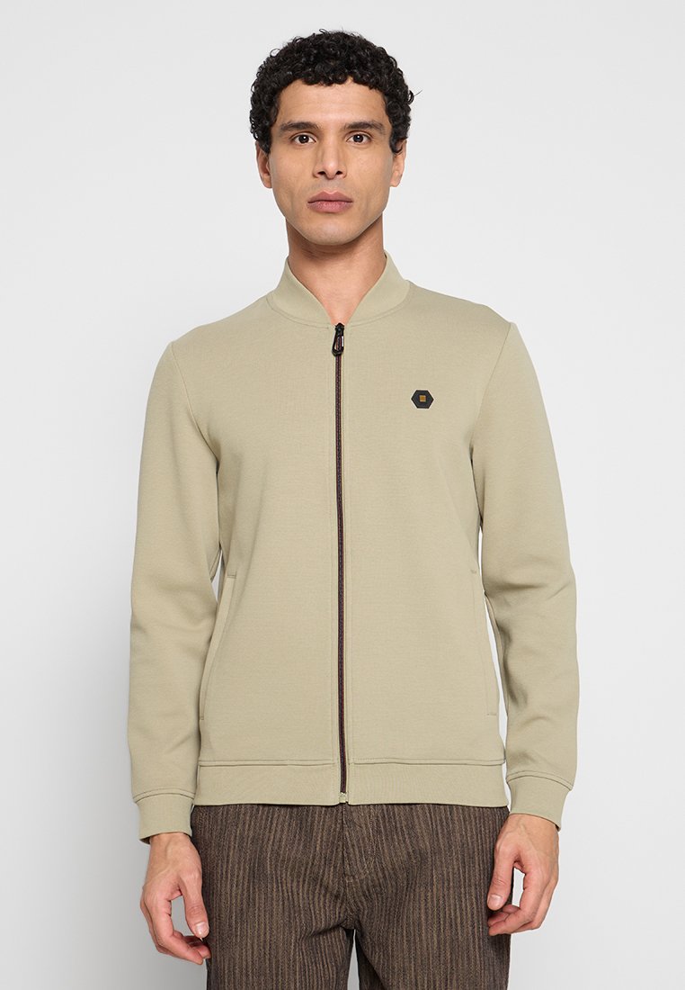 Beige jas met ritssluiting en opstaande kraag, met een klein zwart zeshoekig logo op de borst en twee zijzakken.