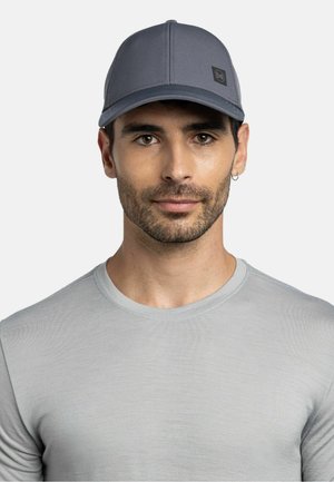 Mann mit kurzen dunklen Haaren und Bart, der eine graue Kappe mit Logo und ein hellgraues Rundhalsshirt trägt, frontal vor einem schlichten Hintergrund.