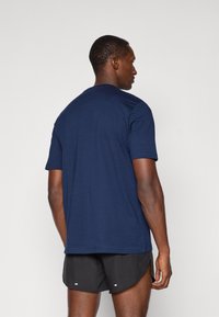 Mand står med ryggen til iført en navy blå T-shirt og sorte sports shorts foran en ensfarvet lys baggrund.