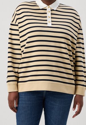 Person iført en beige og sort langærmet skjorte med vandrette striber, hvid krave og guldknapper, kombineret med blå jeans.