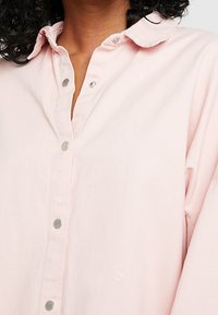 Ljusrosa skjorta med knappar i mjukt tyg, med en spetsig krage och silverfärgade tryckknappar fram.