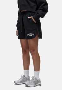 Pantaloni corti neri in cotone con cintura elastica, tasche laterali e testo ricamato bianco. Indossati con scarpe da ginnastica argentate e calze bianche.