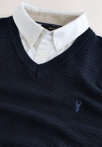 Navy blue textured V-neck sweater layered over a white button-down collar shirt with a small deer logo on the chest.