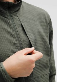 Grön texturerad jacka med dragkedja och hög krage samt sidoficka. Hand som håller i fliken vid dragkedjan, vilket visar material och design.