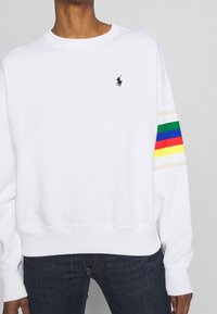Bílá mikina s černým logem, vícebarevnými pruhovanými doplňky na rukávech, kulatým výstřihem a žebrovanými man žetami pro pohodlí.