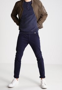 Veste aviateur vert olive, pull à côtes bleu marine, pantalon slim bleu marine et baskets blanches. Détails visibles de fermeture éclair et boutons, ensemble décontracté.