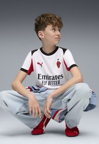 Maglietta da calcio bianca a maniche corte con accenti neri e rossi. Collo nero, maniche rosse, logo anteriore con la scritta "Emirates Fly Better". Jeans blu, scarpe rosse.