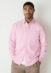 Helles rosa Hemd mit langen Ärmeln, einer Brusttasche und einem strukturierten Stoff. Übereinander mit einem weißen T-Shirt getragen und kombiniert mit beigen Hosen.