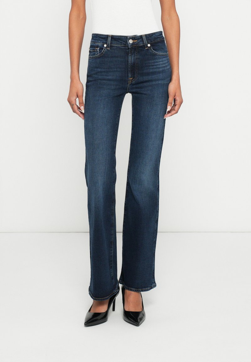 Jeans din denim albastru închis, cu croială dreaptă, estompare subtilă și buzunare frontale. Purtate cu pantofi negri cu toc înalt.