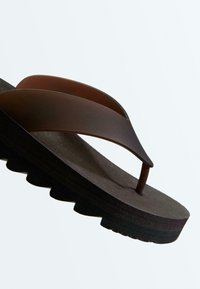 Bruine teenslipper met dikke zwarte geribbelde zool en brede bandjes tegen een effen lichte achtergrond.
