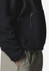 Black fleece jacket with zippered pockets, textured fabric, and a relaxed fit, paired with light-colored pants in a standing pose.