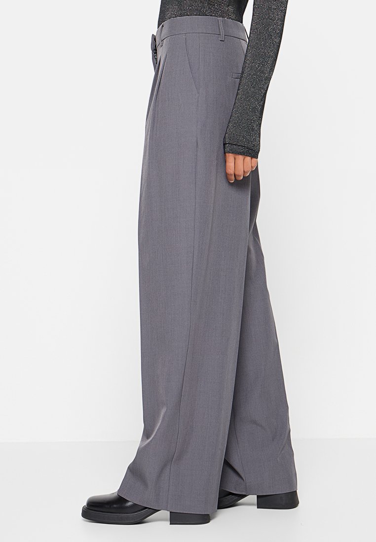 Graue, weit geschnittene Hose aus einem glatten Stoff, mit hoher Taille, seitlichen Taschen und einem maßgeschneiderten Schnitt. Kombiniert mit schwarzem Schuhwerk.