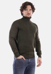 Maglione a collo alto lavorato a maglia di un verde scuro, con un motivo texture, design aderente e polsini a coste, indossato con jeans in denim blu.