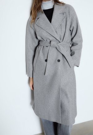 Femme portant un manteau gris en laine à double boutonnage ceinturé, sur un col roulé gris clair et un haut noir, debout devant un fond blanc.