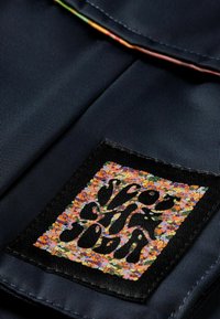 Nadmorska tkanina o teksturowym wykończeniu zawiera kolorową naszywkę z stylizowanym logo z pomarańczowymi, różowymi i zielonymi akcentami na czarnym tle.