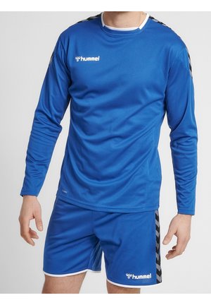 Man draagt blauwe Hummel sportkleding met een top met lange mouwen en korte broek met zwarte chevronstrepen op de schouders en zijkanten.