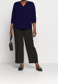 Blusa blu navy a scollo a V con maniche a tre quarti, abbinata a pantaloni neri a gamba larga e scarpe nere con dettagli dorati. Tiene in mano una borsa nera.