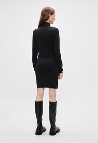 Vestido preto de manga longa com silhueta ajustada e gola alta, combinado com botas pretas até ao joelho com detalhe de fecho lateral.