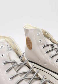 Sneakers high-top beige realizzati in materiale liscio, con occhielli argentati, lacci neri e una toppa con logo marrone di lato.