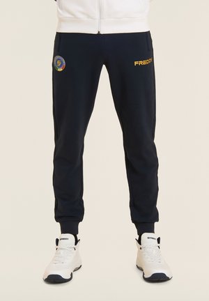 Persona con joggers negros con el logo "FREDDY" y un parche redondo, combinados con zapatillas blancas de caña alta con cordones.