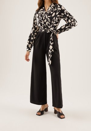 Czarna kopertowa bluzka z białym abstrakcyjnym wzorem w połączeniu z czarnymi spodniami o szerokich nogawkach; modelka ma na sobie czarne sandały na słupku.
