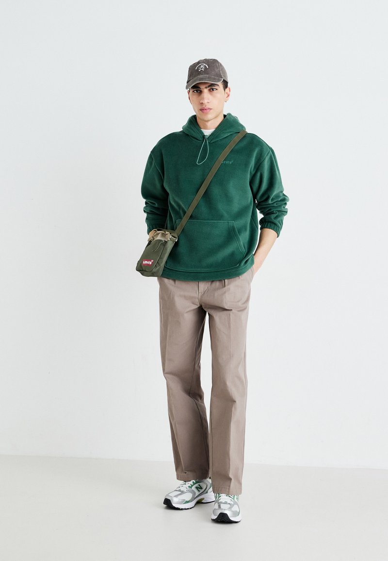 Grüne Fleece-Hoodie, beiges Paar Hosen, graue Mütze, grüne Umhängetasche und graue Sneakers mit weißen und grünen Akzenten. Schlichter Hintergrund.