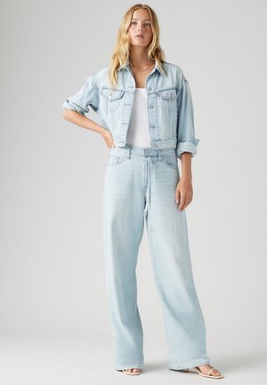 Gai zila džinsa jaka ar saīsinātu dizainu un divām priekšējām kabatām, savienota ar plašām gaiši zilām džinsa biksēm un baltu topu.