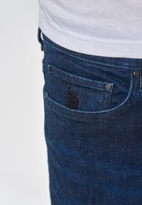 Zilu denim bikses ir ar aizmugures kabatu, kas rotāta ar tumšu izšuvumu un sudraba aksesuāriem. Tekstūra izskatās гладка ar nelielu izbalēšanu.