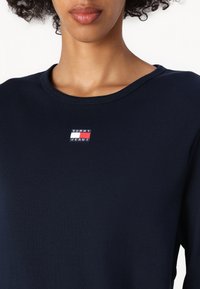 Jūros mėlynos spalvos raštuotas ilgomis rankovėmis viršus su plačiu iškirpimu ir mažuoju siuvinėtu Tommy Jeans logotipu raudonos, baltos ir mėlynos spalvų ant krūtinės.