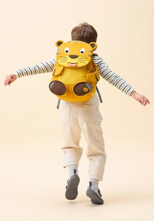 Child wearing striped shirt and light pants with arms outstretched, carrying yellow tiger-shaped backpack, walking away on a beige background.