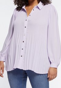 Lila plisserad blus med knäppning fram, långa ärmar och rund nederkant. Har silverknappar som accent och är tillverkad av mjukt, lätt tyg.