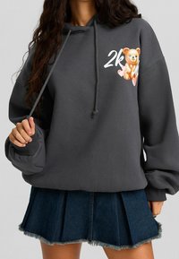 Sudadera gris de gran tamaño con cordones, que presenta un gráfico de un oso de dibujos animados y el texto "2K". Combinada con una falda de mezclilla con pliegues y dobladillo deshilachado.