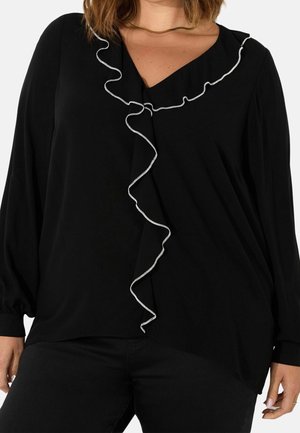 Femme portant un chemisier noir à manches longues avec un volant bordé de blanc courant verticalement du col à l'ourlet.