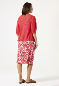 Pull en maille rouge �à manches trois-quarts associé à une jupe imprimée longue jusqu'aux genoux en rose et blanc, et des sandales à glisser brunes.