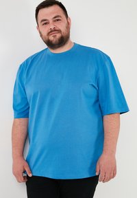 Modrá tričko s krátkym rukávom vyrobené z mäkkého materiálu. Má okrúhly výstrih a voľný strih, nosené cez čierne nohavice na jednofarebnom pozadí.