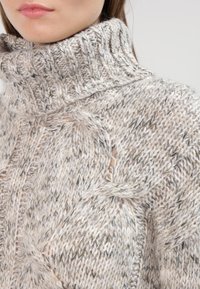 Pull en maille col roulé beige clair avec motifs texturés. Présente un col haut et de légères ouvertures dans le design pour plus de détails.