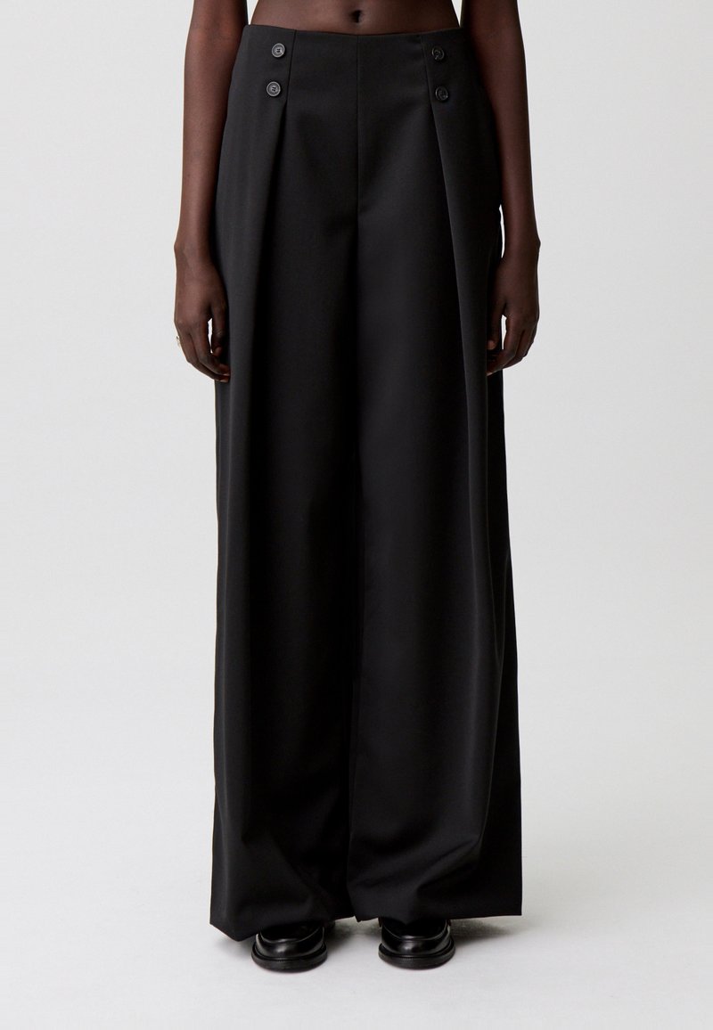 Pantalones negros de pierna ancha hechos de tela texturizada, con pliegues frontales y dos botones decorativos en la cintura. Acabado liso, corte entallado.