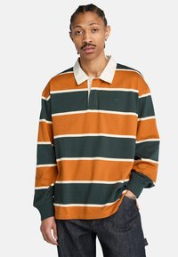 Mężczyzna z warkoczami nosi zieloną i pomarańczową szeroko paskowaną koszulkę rugby z białym kołnierzykiem, stojąc z jedną ręką w kieszeni dżinsów.