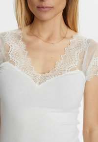 Top de tirantes blanco con canalé y escote en forma de V, adornado con intrincados detalles de encaje crema en los hombros y el escote. Diseño sencillo y ajustado.