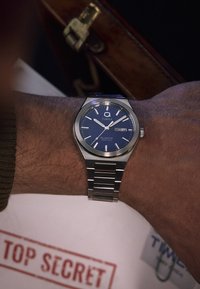 Ceas din oțel inoxidabil argintiu cu un cadran albastru, indicatori de oră argintii și un display pentru dată. Brățară de tip link.